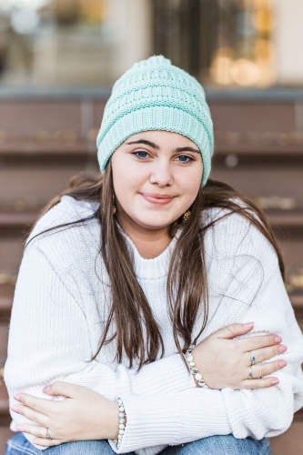 Teenager sitting on steps leaning forward with arms crossed smiling wearing beanie - Australian Stock Image