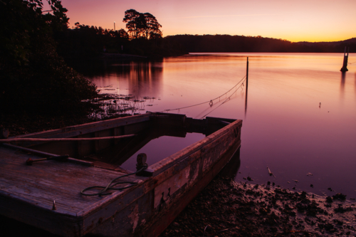The idyllic setting across Wagonga Inlet and a disused boat at sunset in Narooma, NSW, Australia - Australian Stock Image