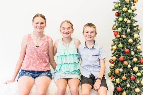 Three children brother and sisters sitting arm in arm on rug next to Christmas tree