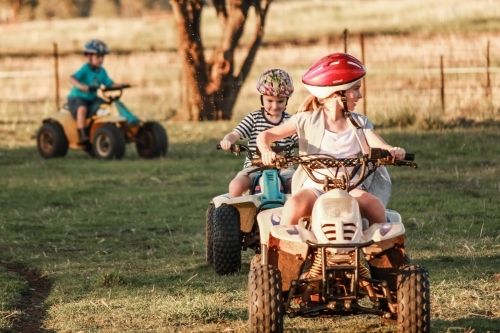 Three children riding on quad bikes around farm property - Australian Stock Image