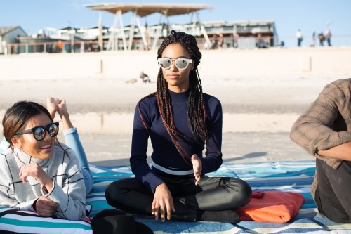 Three friends sitting on a picnic blanket relaxing at the beach - Australian Stock Image
