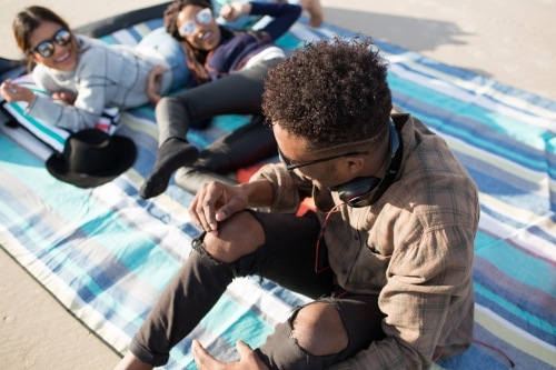 Three friends sitting on a picnic blanket relaxing at the beach - Australian Stock Image