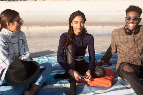 Three happy friends sitting on a picnic blanket at the beach - Australian Stock Image