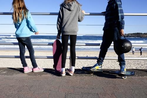 Three kids standing with skateboards looking out at the beach - Australian Stock Image