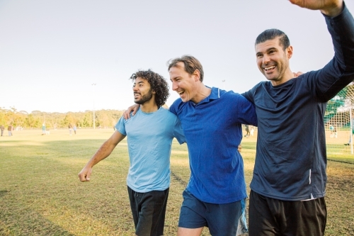 Three men walking together on a soccer field, laughing and smiling, celebrating a win - Australian Stock Image