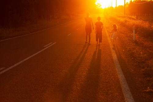 Three people walking along the roadside towards the sunset in golden light - Australian Stock Image