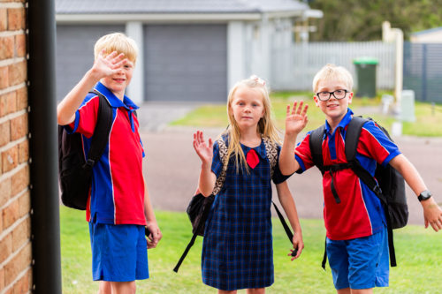 Three siblings ready for school waving goodbye at the door of home  - Australian Stock Image