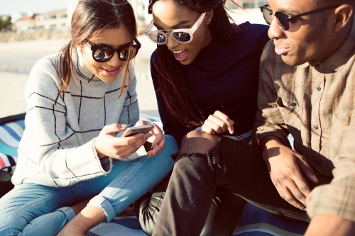 Three teenage friends sitting looking at a phone on a picnic rug - Australian Stock Image