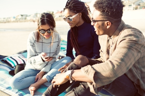 Three teenage friends sitting looking at a phone on a picnic rug - Australian Stock Image