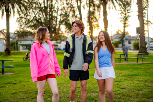 Three young adults leisurely stroll through a park as the sun sets behind palm trees - Australian Stock Image