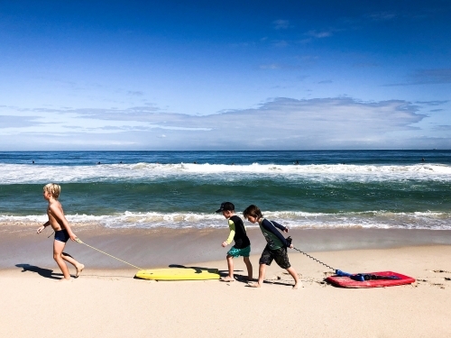 Three young boys dragging boogie boards along shoreline of beach