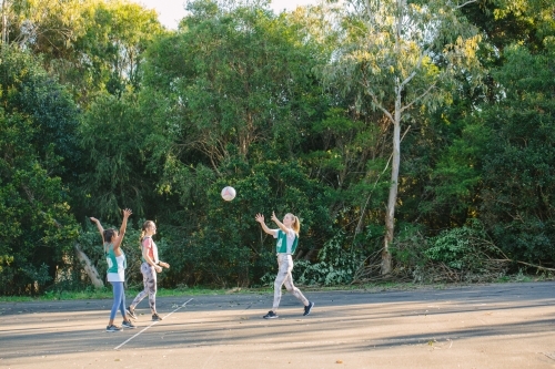 three young women with one throwing a net ball in mid air on a sunny day - Australian Stock Image