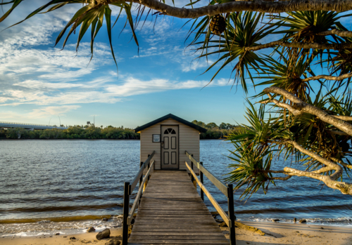Timber boat shed and jetty on sandy river shoreline with pandanus tree, blue sky and clouds - Australian Stock Image