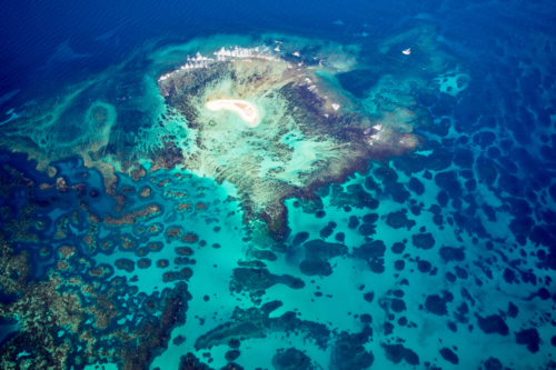 Tiny coral island surrounded by vivid turquoise reef and deep ocean blue. - Australian Stock Image