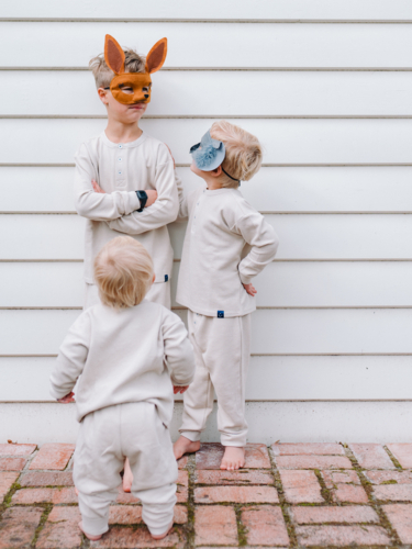 Toddler boy walking towards older brothers wearing animal masks standing in front white walls - Australian Stock Image