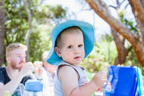 Toddler boy with a blue brim hat, sits on table enjoying picnic with family.