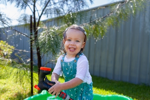 Toddler girl in suburban backyard playing with hose and getting wet in summertime - Australian Stock Image