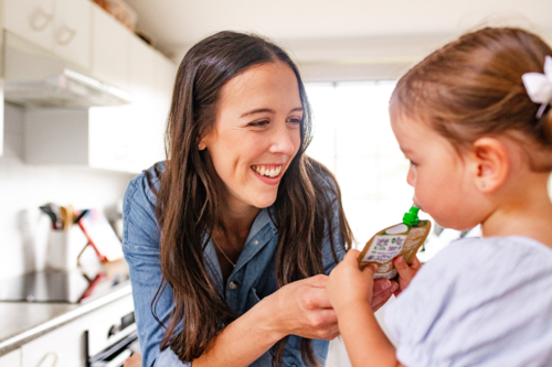 Toddler girl standing sipping from food pouch in the kitchen - Australian Stock Image