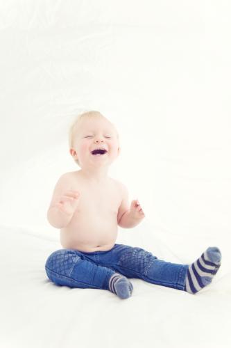 Toddler sitting on bed under the sheets wearing jeans and socks laughing - Australian Stock Image