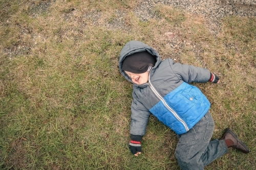 Top down view of little boy lying on grass in winter with cheeky smile - Australian Stock Image