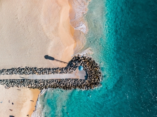 top shot of a beach front with a small walkway made of rocks, lamp post and white sand shoreline - Australian Stock Image