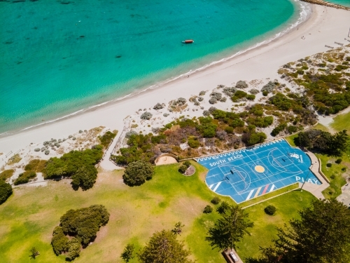 top view shot of a blue basketball court near a beach with white sand, grass plains and green bushes