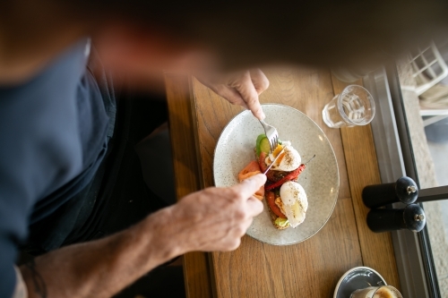 Top view shot of a man slicing a toast with avocado, poached eggs, salmon on a plate
