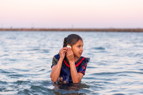 Torres Strait Islander child listening to shell in ocean water at dusk - Australian Stock Image