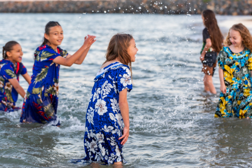 Torres Strait Islander girls in traditional floral dresses playing and splashing in ocean water - Australian Stock Image