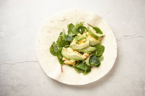 Tortilla wrap on a white surface topped with avocado slices, spinach, and scrambled egg - Australian Stock Image