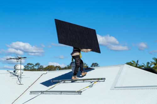 Tradesman carrying solar panel onto metal roof of suburban house - Australian Stock Image