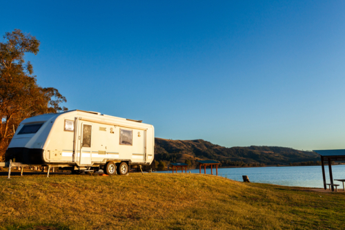 Travel concept – caravan parked by lakeside in sunset light with blue sky copy space - Australian Stock Image