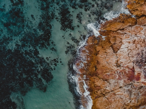 Turquoise blue water crashing against orange rocky shore – abstract aerial - Australian Stock Image