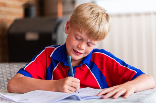 Tween boy in school uniform doing bookwork homework on table in backyard at home - Australian Stock Image