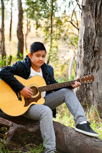 Tween boy playing guitar in bushland seated on log