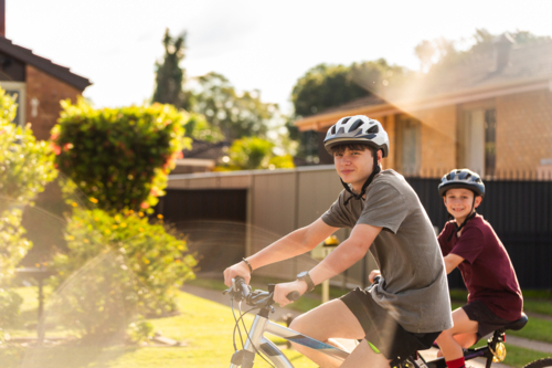 Two Australian kids riding push bikes together on sunlit street in country town - Australian Stock Image