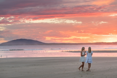 Two blonde girls in white summer dresses on a beach at sunset