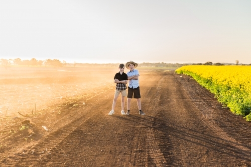 Two boys standing close together arms crossed on dusty dirt road on farm next to canola field