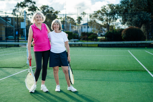 Two elderly women stand confidently on a tennis court, holding racquets and smiling warmly. - Australian Stock Image