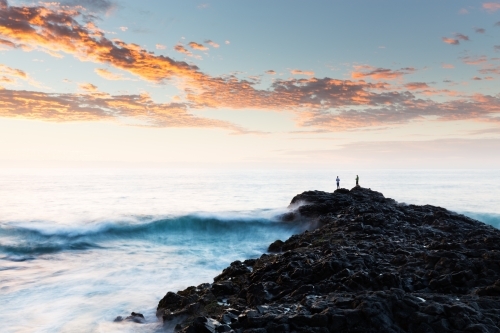 Two fishermen fish in the Pacific Ocean from a rocky headland in a long exposure sunrise scene - Australian Stock Image