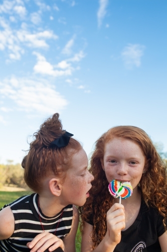 Two girls outside eating a rainbow lollipop - Australian Stock Image