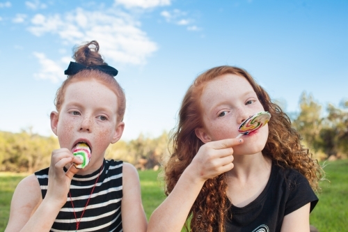 Two girls outside eating rainbow lollipops - Australian Stock Image
