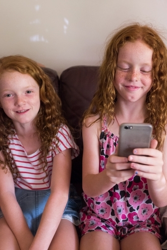 Two girls sitting on a couch playing on a smartphone - Australian Stock Image