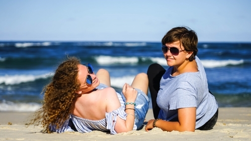 Two girls wearing sunglasses sitting on the sand - Australian Stock Image