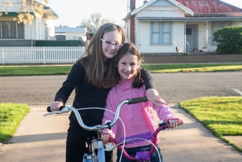 Two happy sisters posing out the front of their house on their bikes. - Australian Stock Image