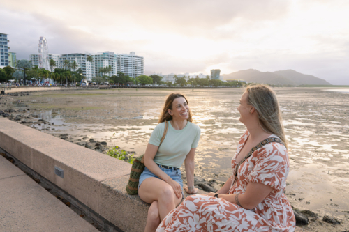 Two happy women talking side by side on a stone ledge overlooking the coastal landscape at sunset - Australian Stock Image