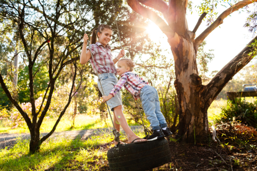 Two happy young Australian children swinging together in front yard of rural home on tyre swing - Australian Stock Image