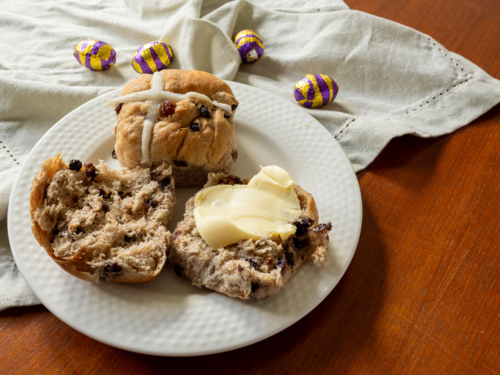 Two hot cross buns, one with butter, on white plate with linen napkin and foil eggs - Australian Stock Image