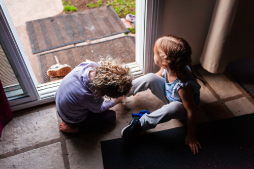 two little girls by back door of home helping each other get shoes on - Australian Stock Image