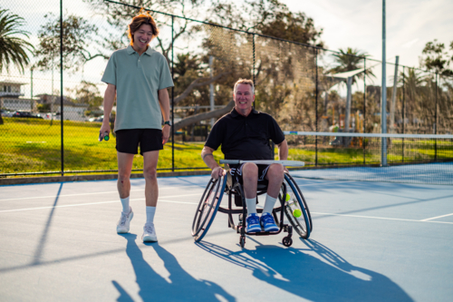 Two men, one in a wheelchair, entering a tennis court in a sunny afternoon - Australian Stock Image